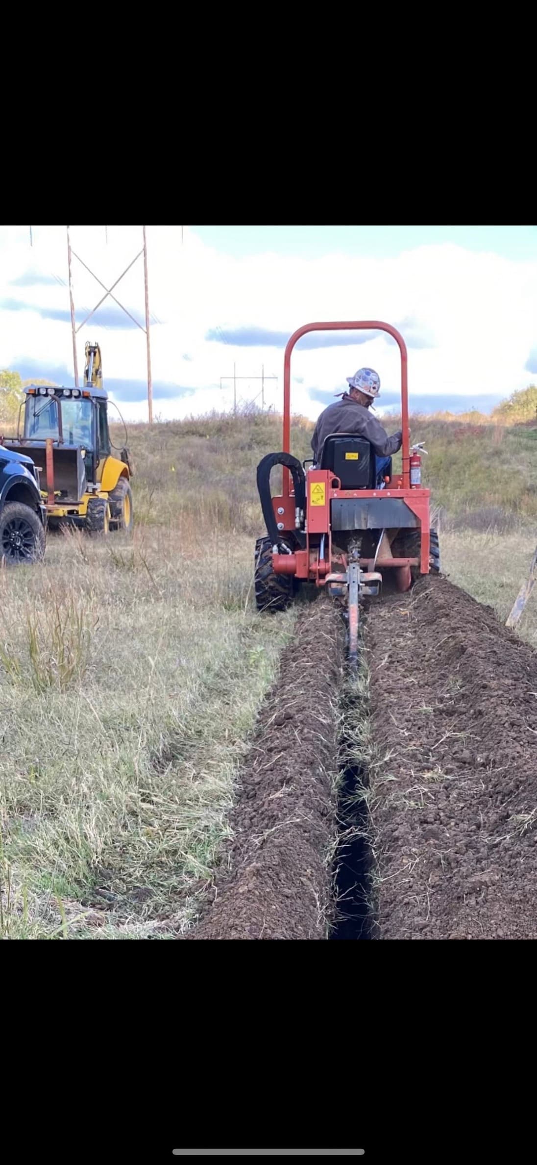 A ride-on trencher cutting a narrow trench across a field, with a backhoe and pickup behind it