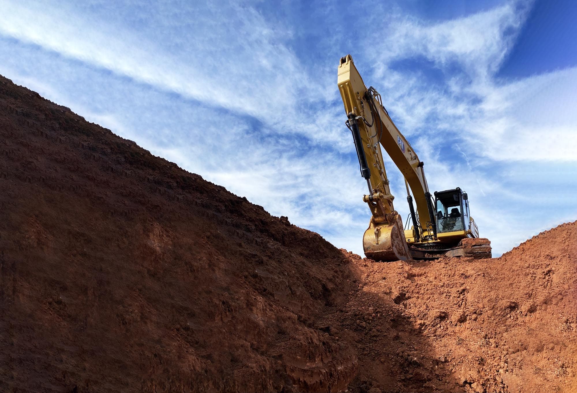 A tracked excavator parked at the crest of a freshly cut bank of red earth under a blue sky