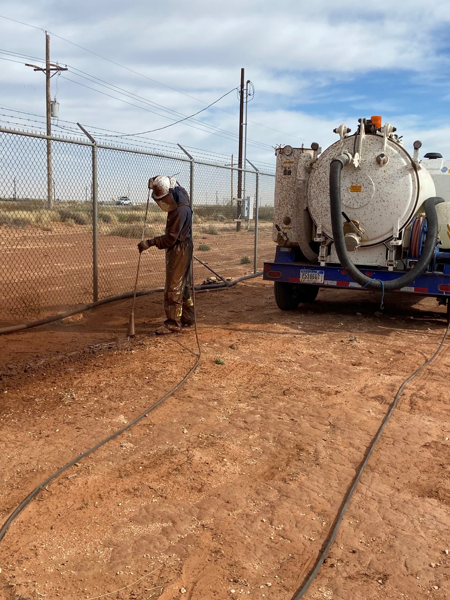 A crew member in coveralls working a hydro-excavation wand beside a vacuum truck at a fenced facility