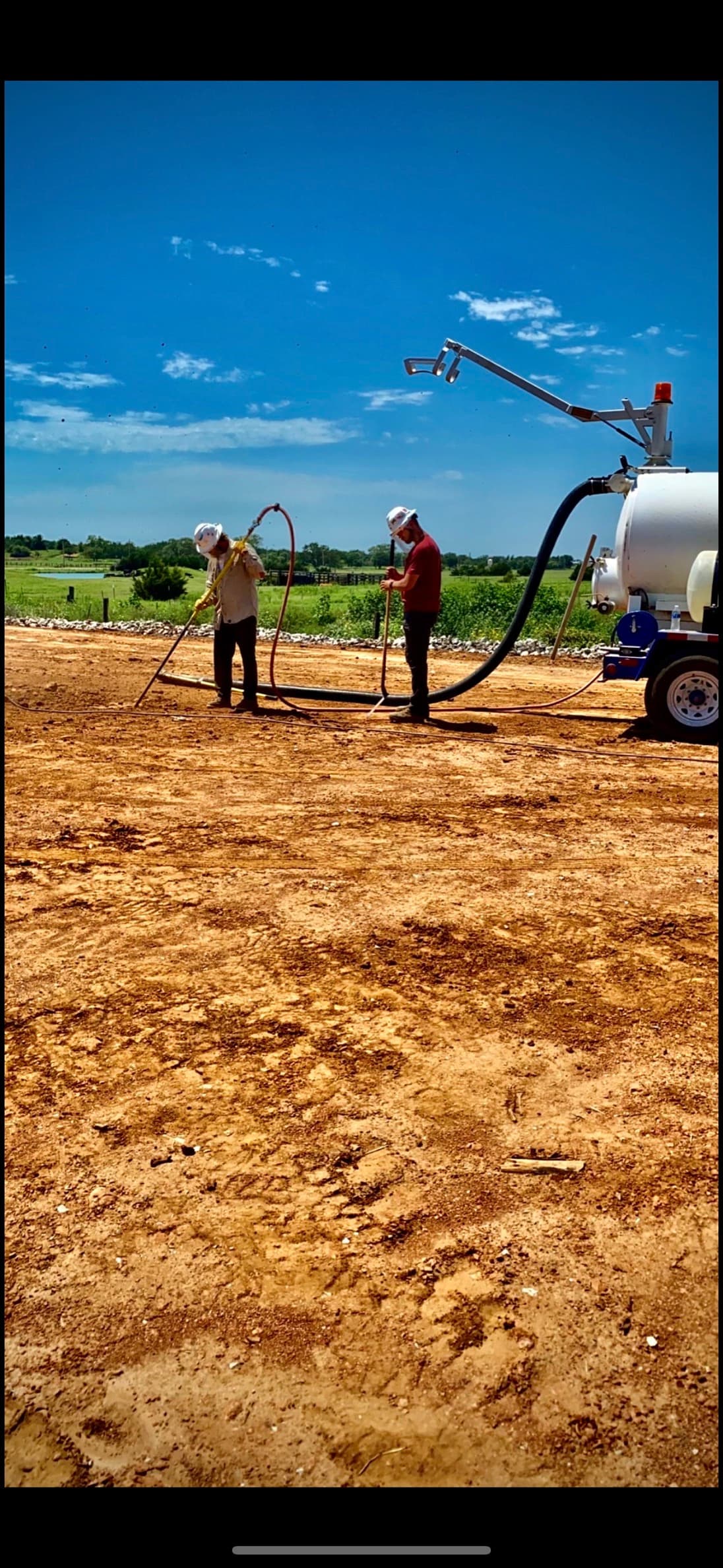 Two crew members working hydro-excavation wands on a graded pad beside a white vacuum trailer